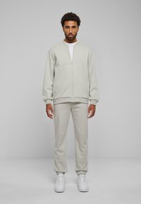 Giacca grigia chiara con zip e pantaloni joggers abbinati realizzati in tessuto morbido. Le scarpe sono bianche. Design semplice e rilassato con polsini e caviglie a coste.