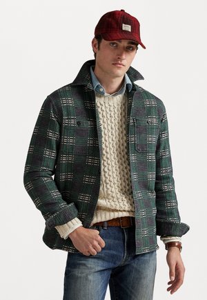 Jauns vīrietis ar sarkanu šalli, zaļu un pelēku pārbaudes jaku virs krēmkrāsas adītā džempera, zilu džinsiem un brūnu ādas pulksteņu.