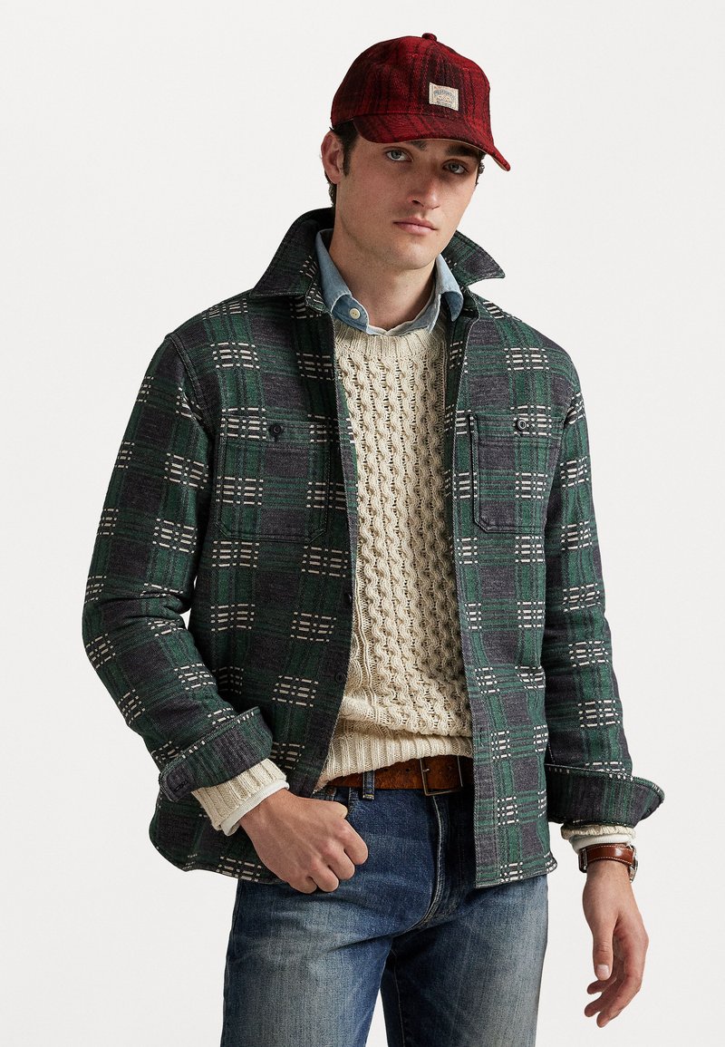 Mladý muž nosi červenú károvanú čiapku, zeleno-sivú károvanú bundu cez krémový sveter s pleteninou, modré džínsy a hnedé kožené hodinky.
