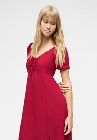 Ξανθιά γυναίκα με μακριά μαλλιά που φορά κόκκινο φόρεμα με κοντά μανίκια, κοιτάζοντας προς τα κάτω και πλάγια, στέκεται μπροστά σε ένα απλό φωτεινό φόντο.