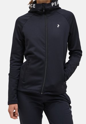 Kobieta w czarnej kurtce na suwak i dopasowanych spodniach, kołnierz kurtki z napisem "PEACE", ręce w kieszeniach na jednolitym tle.