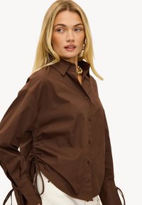 Chemise marron à boutons avec un col pointu, comportant des côtés froncés et des liens ajustables. Confectionnée en tissu lisse, avec des manches longues.