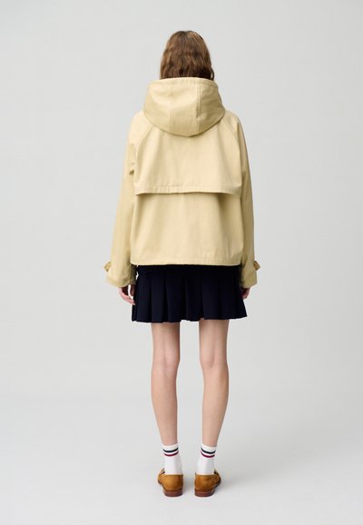 Veste à capuche beige au design court, dotée d'un rabat à l'arrière, associée à une jupe plissée bleu marine et des chaussures marron. Chaussettes blanches avec des rayures.