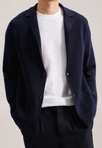 Marinblå stickad kavaj med slagkrage, två knappar och fickor med lappar, över en vit t-shirt med rund halsringning och mörka byxor.