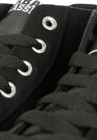 Must musta tossud, millel on metallist nööriaugud, mustad nöörid ja tekstuuriga kangas. Kinga keele osal on valge logo.