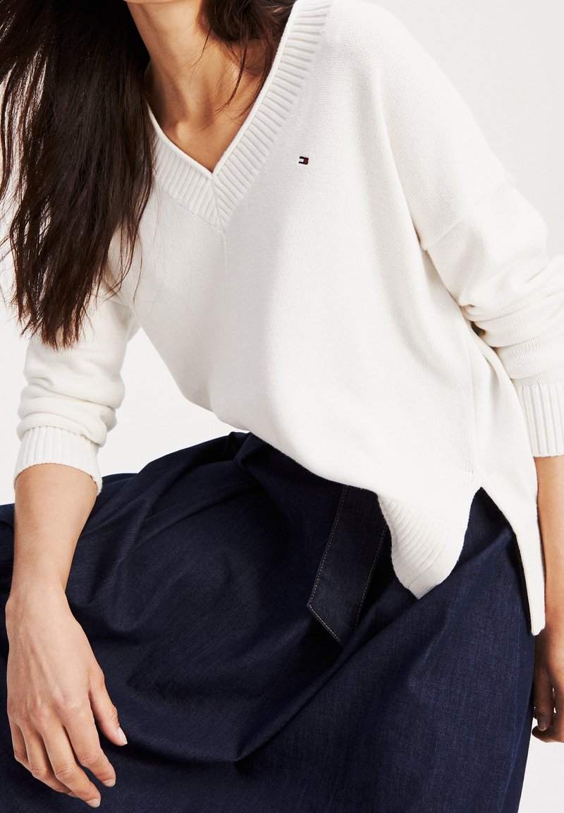 Femme portant un pull blanc à col en V avec des poignets côtelés et une jupe bleu marine, assise avec une main reposant sur son genou.