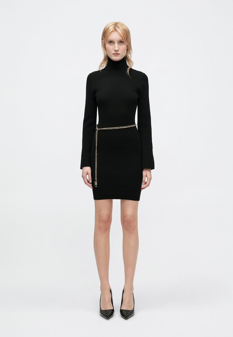 Fekete bordázott ruha magas nyakkivágással és hosszú ujjakkal, arany lánccal megkötve a deréknál. Fekete hegyes sarkú cipővel viselve.