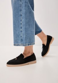 Black suede loafers with a flat beige rubber sole. Features a rounded toe and a decorative strap with hardware detail. Worn with cropped denim.