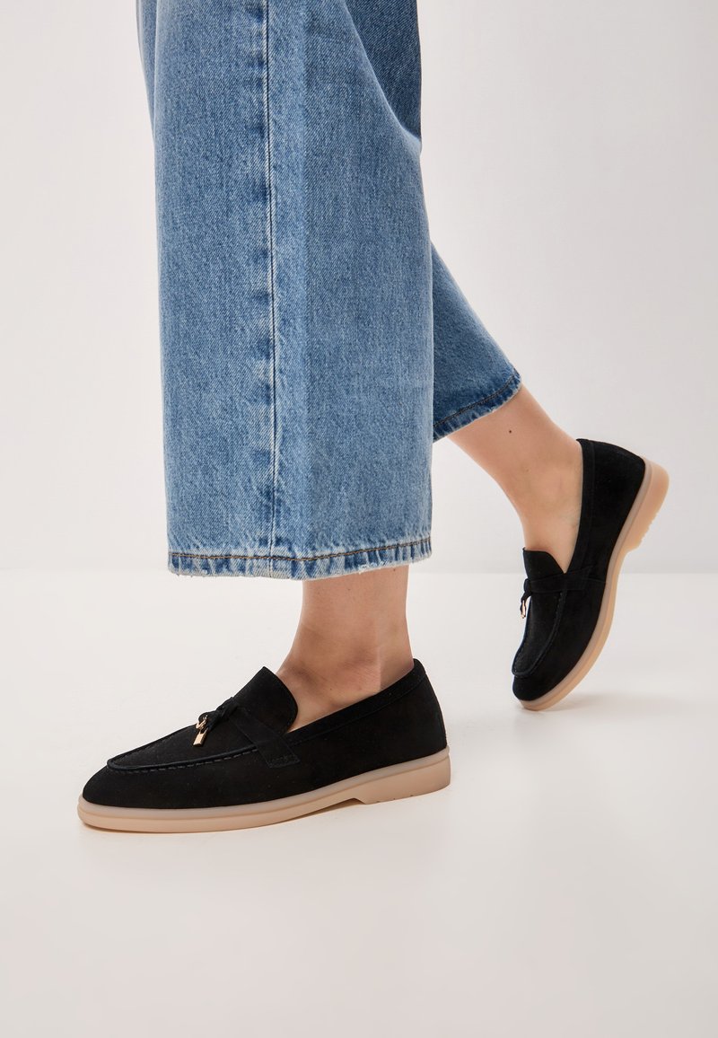 Black suede loafers with a flat beige rubber sole. Features a rounded toe and a decorative strap with hardware detail. Worn with cropped denim.