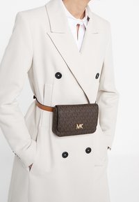 Nainen, jolla on päällään valkoinen kaksirivinen takki mustilla nappeilla ja ruskea Michael Kors -vyölaukku vyön ympärillä.