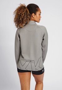 Grå, langærmet atletisk top med høj krave og sort geometrisk mønster på ryggen. Glat tekstur, tætsiddende design.