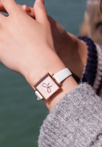 Reloj cuadrado con una caja de oro rosado, correa de cuero blanco y una esfera rosa con un diseño decorativo de cinta y acentos de cristal.