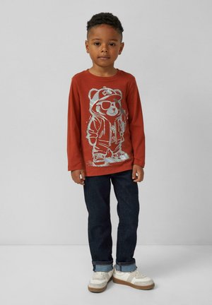 Jongen staat tegen een grijze achtergrond gekleed in een roestkleurig langarmshirt met een opdruk van een beer in zonnebril, donkere spijkerbroek en witte sneakers.