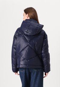 Jūras spēku puffer jaka ar dimanta kviltiem, pārliecinošu kapuci un elastīgām piedurknēm, savienota ar vaļīgām džinsa biksēm.