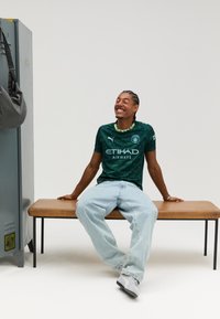 Groen voetbalshirt met een geometrisch patroon, voorzien van witte merktekens en accenten. Gecombineerd met lichtblauwe wijde spijkerbroek en grijze schoenen.