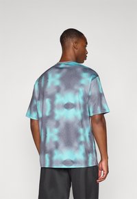 T-shirt med korte ærmer, der har et tie-dye mønster i nuancer af teal og grå. Glat tekstur, rund hals og afslappet pasform. Bæres med sorte shorts.
