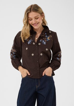 Kvinde smilende med hænderne i lommerne, iført en mørk skjorte med blå blomsterbroderi og mørke jeans mod en ensfarvet lys baggrund.