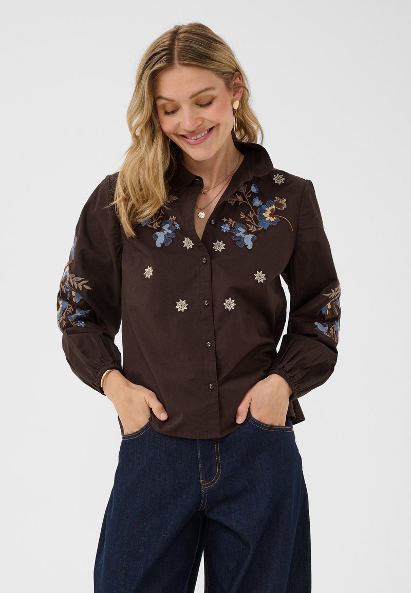 Femme souriante avec les mains dans les poches, portant une chemise sombre à broderie florale bleue et un jean foncé sur un fond uni clair.