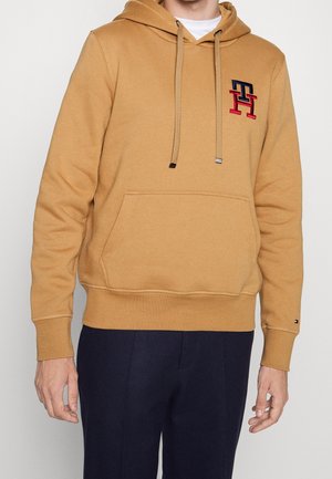 Hombre vistiendo una sudadera color camel con bolsillo frontal y pantalones azul marino, logo bordado en el pecho y pequeño detalle de bandera en el puño de la manga.
