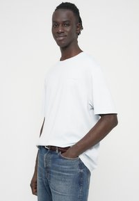 Ανοιχτόμπλε βαμβακερό T-shirt με κοντά μανίκια, στρογγυλή λαιμόκοψη και διακριτικό λογότυπο στο στήθος, συνδυασμένο με ξεθωριασμένο μπλε τζιν και υφαντή ζώνη.