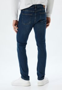 Jeans de mezclilla azules con un ajuste slim, que cuentan con dos bolsillos traseros, detalles de costura sutiles, combinados con zapatillas blancas. Textura limpia y suave.