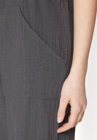 Gray fabric garment with a textured surface, featuring a side pocket and a fitted silhouette, with a hand positioned near the pocket.