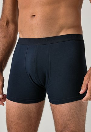 Mand iført sorte tætsiddende boksershorts med brandet taljebånd, der viser overkroppen og det øverste af benene mod en ensfarvet baggrund.