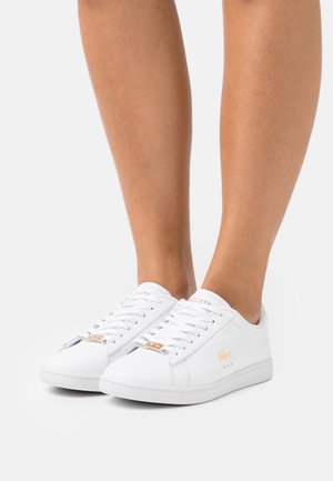Zapatillas blancas de cuero con punteras redondas, suelas de goma planas, diseño con cordones y detalles en tono dorado con el logo de Lacoste.