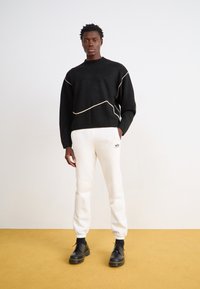 Sort sweater med hvidt linje-design, parret med cremede sweatpants med logo. Model bærer sorte snørebåndsstøvler og står på en gul overflade.