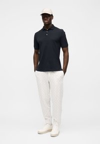 Marineblaues Poloshirt, weiße gemusterte Hose mit strukturierendem Design, hellfarbige Mütze und Turnschuhe. Einfaches, lässiges Outfit an einem stehenden Modell.