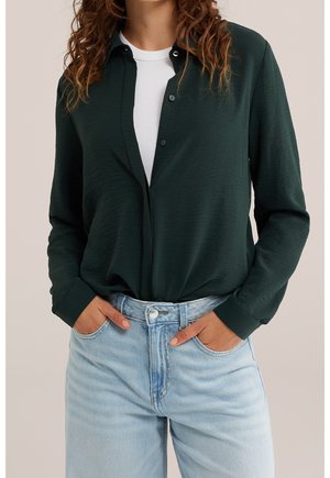 Vrouw die een donkergroene blouse draagt over een wit topje en lichtblauwe spijkerbroek met haar handen in haar zakken, bijgesneden ter hoogte van de schouders en heupen.