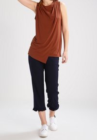 Braunes ärmelloses Oberteil mit drapiertem Vorderteil, kombiniert mit marineblauen, cropped Hosen mit Rüschensaum. Weiße Sneakers runden den Look ab.