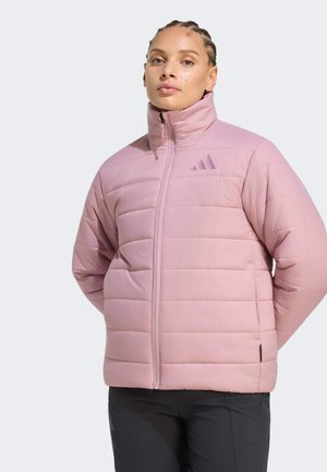 Persoon met gevlochten haar die een lichtroze doorgestikte jas met hoge kraag en zwarte broek draagt, staand tegen een effen achtergrond.