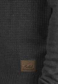 Maglione lavorato a maglia grigio scuro con un motivo testurizzato e un'etichetta rettangolare in pelle marrone con la scritta "Solid" e "United Traditions" in rilievo.