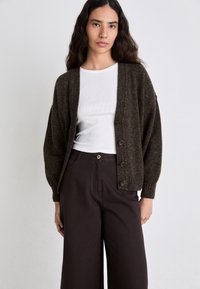 Cardigan marrone lavorato a maglia con maniche oversize, dotato di cinque bottoni. Indossato sopra una camicia bianca aderente e abbinato a pantaloni larghi marrone scuro.