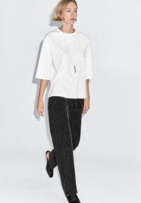 Woman with short blonde hair walking, wearing a loose white shirt, long necklace, black pants, and black shoes against a plain background.