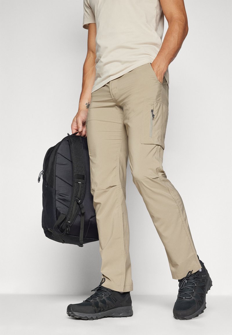 Pantalons de randonnée beiges avec poche latérale et fermeture éclair, associés à un t-shirt beige clair, un sac à dos noir et des chaussures de randonnée noires.