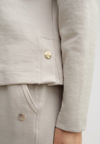 Beige sweatshirt with long sleeves, featuring a gold metal button detail and seamless stitching; paired with matching joggers with a logo accent.
