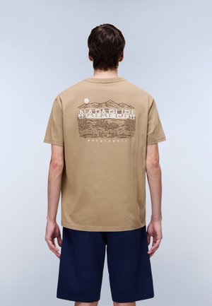 Persoon met rug naar voren draagt beige t-shirt met berg- en natuurelementen en de tekst "NAPAPIJRI geographic", en navy blauwe korte broek.