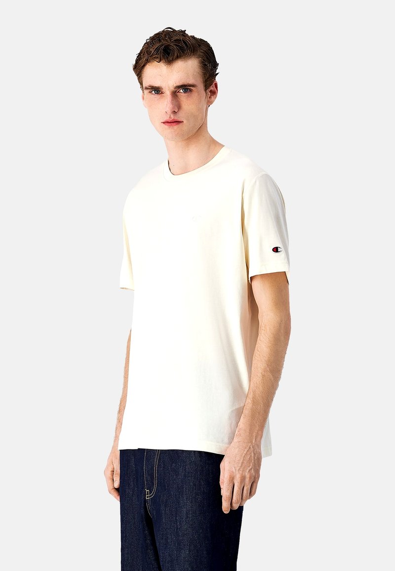 Cremefarbenes T-Shirt mit kurzen Ärmeln aus Baumwolle, mit Rundhalsausschnitt und kleinem Logo auf dem linken Ärmel; kombiniert mit dunkelblauen Jeans.