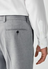 Grijze broek van gestructureerde stof met een achterzak met een zwarte knoop. Een wit overhemd met lange mouwen is gedeeltelijk zichtbaar.