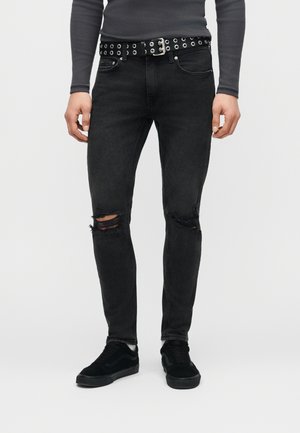 Άτομο που φοράει σκισμένο μαύρο στενό τζιν, μαύρη ζώνη με τρύπες, σκούρο γκρι μακρυμάνικο πουκάμισο και μαύρα αθλητικά, στέκεται μπροστά σε λευκό φόντο.