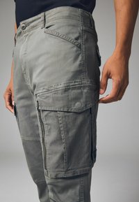 Pantalones cargo de tela gris, con múltiples bolsillos, una cintura ajustada y texturas contrastantes en las piernas. Detalle de cremallera visible.