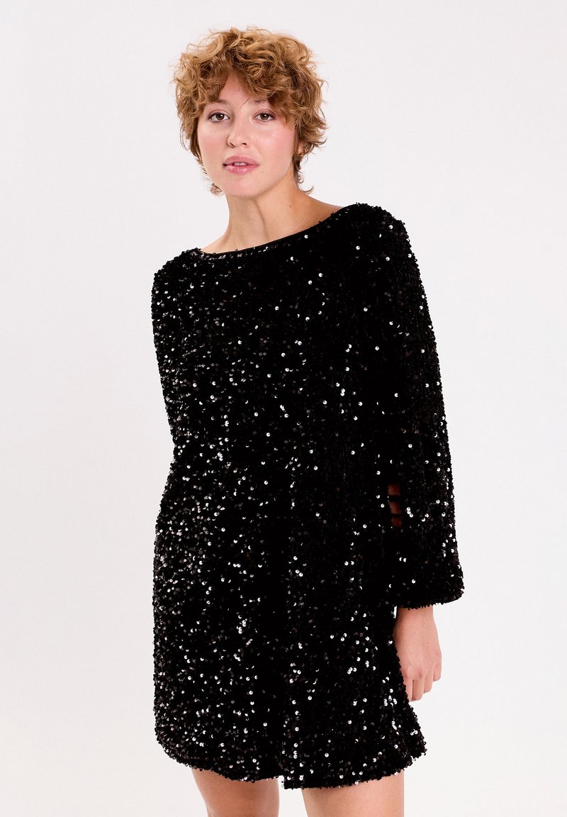 Robe en sequins noire avec une coupe décontractée et des manches longues, dotée d'un décolleté bateau et d'accents ouverts sur les côtés. Texture brillante sur l'ensemble.