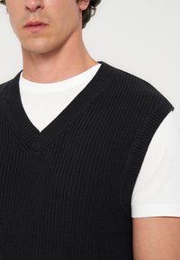Jeune homme portant un pull sans manches en maille noire à col en V par-dessus un t-shirt blanc à manches courtes sur un fond clair.