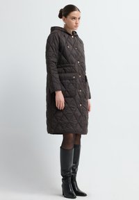 Bruine gewatteerde jas met een capuchon, voorzien van ruitpatronen, drukknoopsluiting en grote zakken. Zwarte laarzen tot de knie complementeren de outfit.