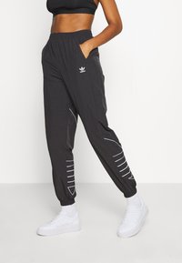 Femme portant un pantalon de survêtement noir Adidas avec des logos et des détails en blanc, associé à des chaussettes blanches et des sneakers blanches, posant avec une main dans la poche.