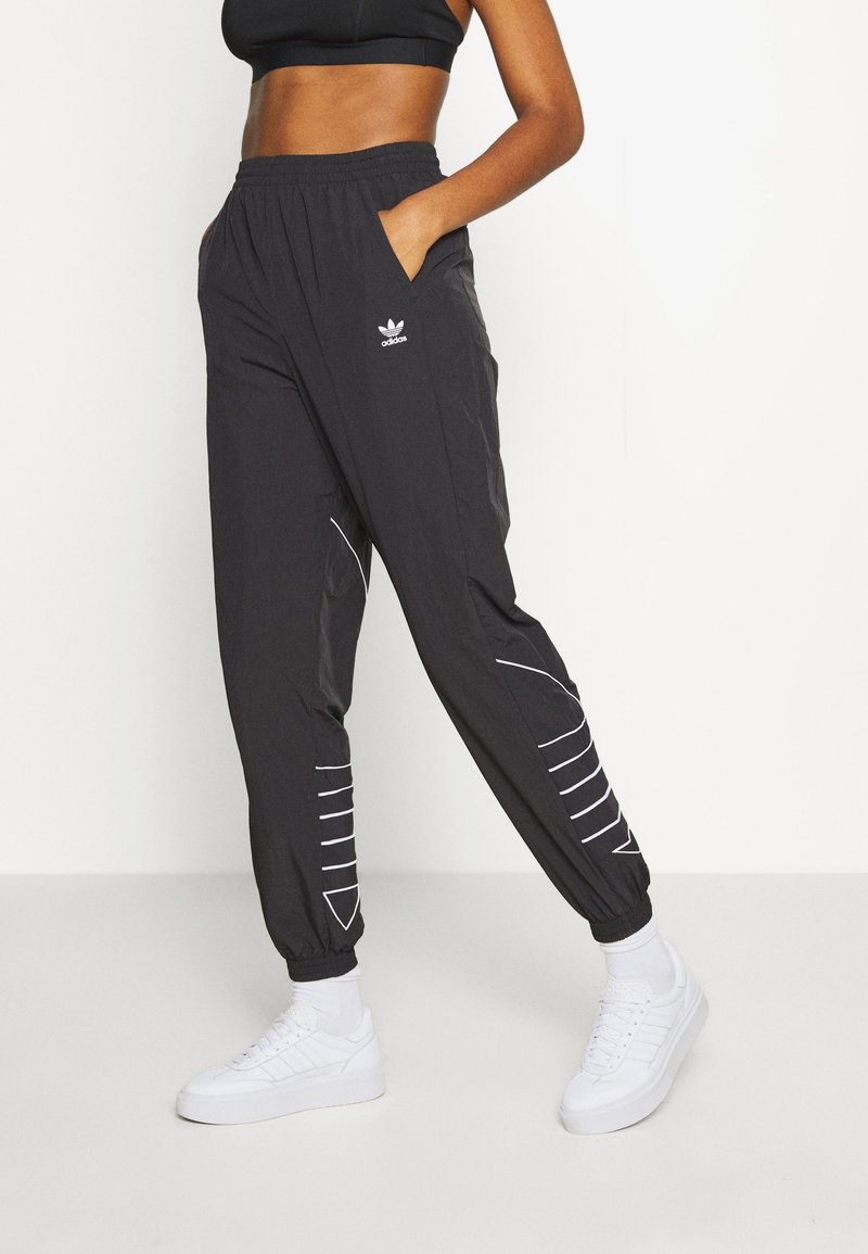 Femme portant un pantalon de survêtement noir Adidas avec des logos et des détails en blanc, associé à des chaussettes blanches et des sneakers blanches, posant avec une main dans la poche.