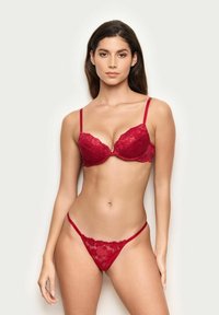 Femme portant un ensemble assorti de lingerie en dentelle rouge, se tenant devant un fond clair et uni, faisant face à l'avant avec une expression neutre.