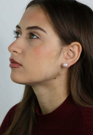 Młoda kobieta z długimi brązowymi włosami, nosząca perłowe kolczyki wkrętki i bordowy golf, patrząca zamyślona w lewo na jednolitym tle.